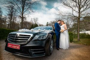 Newlywed couple kissing beside a black Mercedes-Benz with a Just Married sign at an outdoor wedding venue.