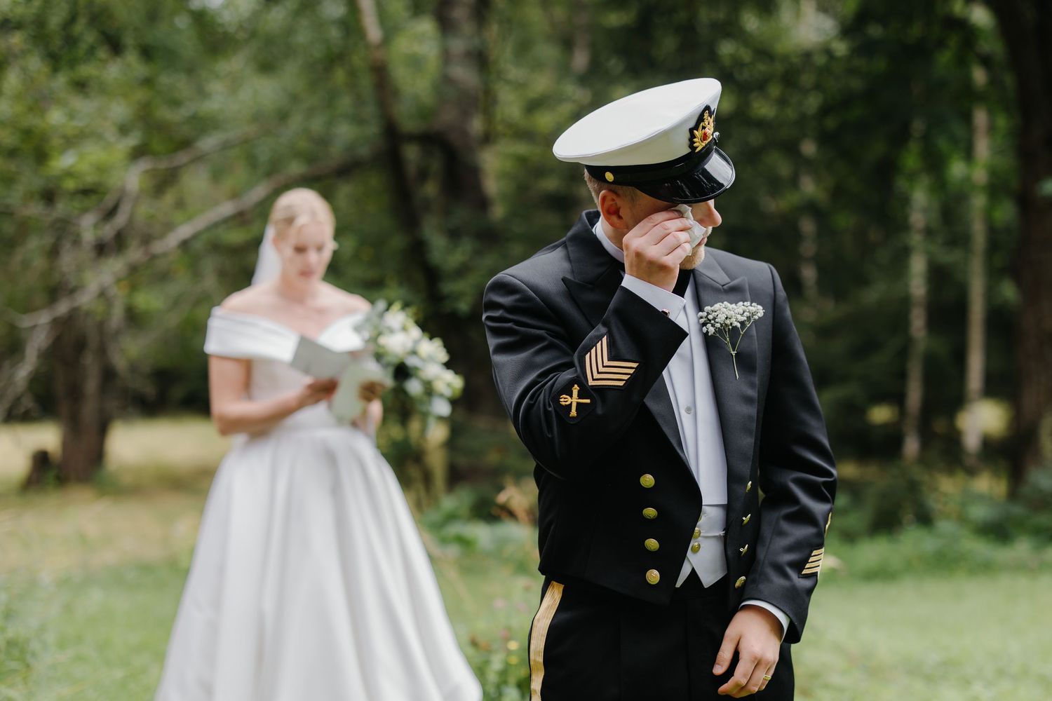Emotional groom in naval uniform wiping tears as bride in white gown stands behind him outdoors.
