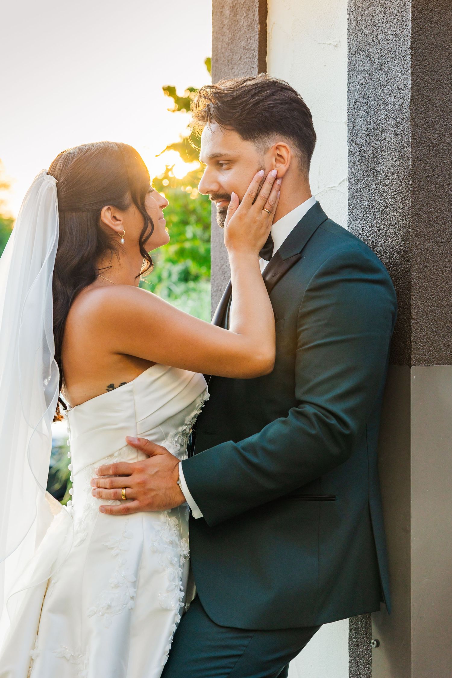 Bride in white gown touches groom's face tenderly as they share a romantic moment outdoors in golden sunset light.