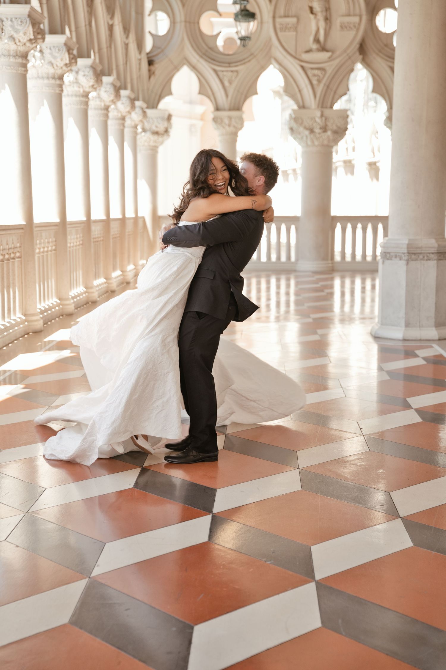 A groom lifts his bride in a white gown, dancing joyfully on a checkered marble floor amid elegant white columns.