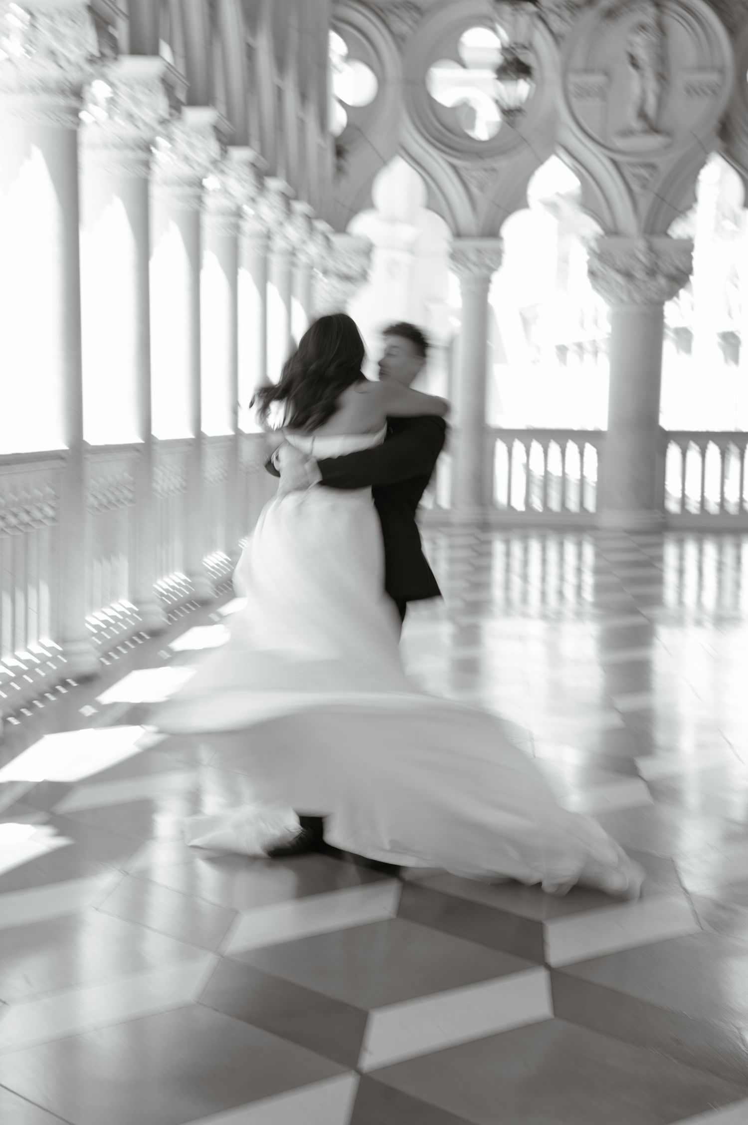 Black and white photo of bride and groom dancing in an ornate columned hall with checkered marble floors.