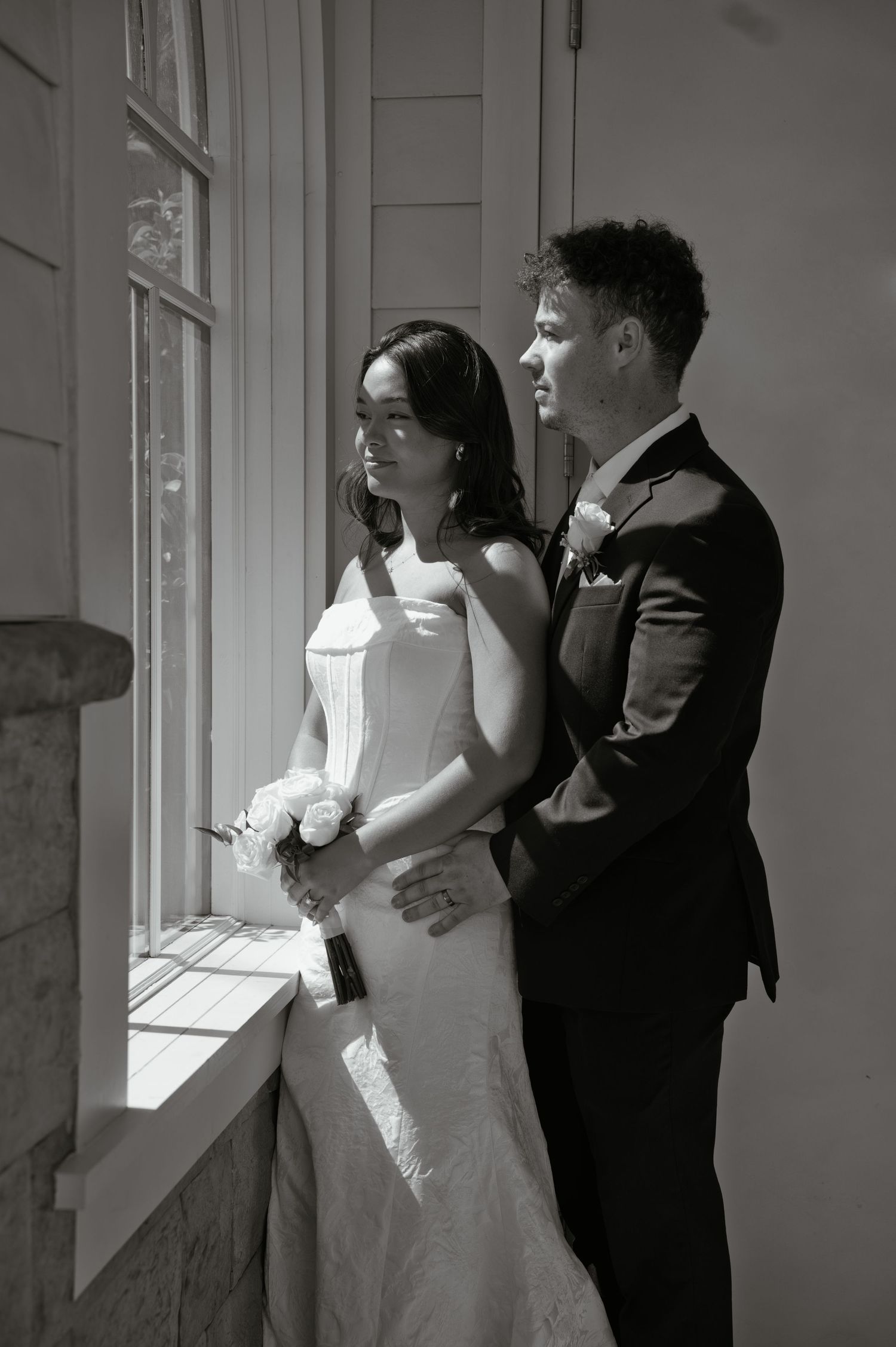 Black and white photo of a bride and groom standing by a sunlit window, gazing outside on their wedding day.