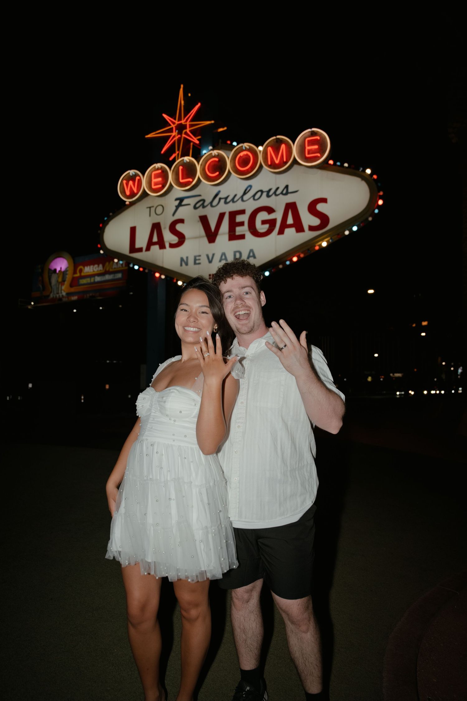 Couple posing and showing off rings in front of the Welcome to Fabulous Las Vegas Nevada sign at night.