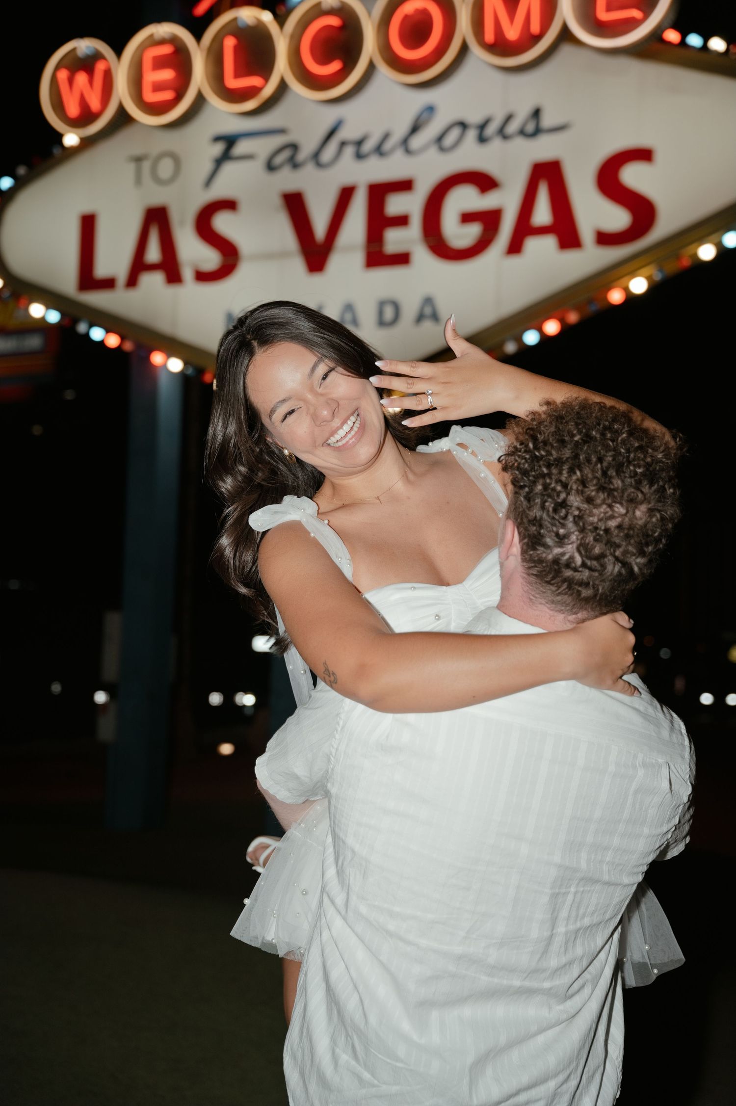 Couple celebrating engagement at the Welcome to Fabulous Las Vegas sign at night, woman showing ring.