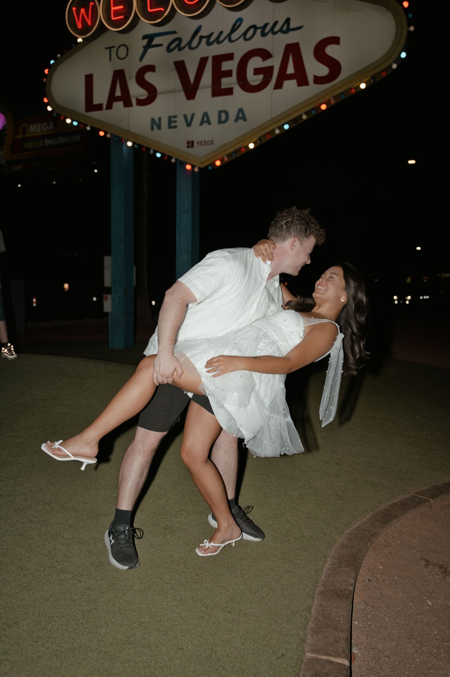 Couple celebrating in front of the Welcome to Fabulous Las Vegas Nevada sign at night, man lifting woman in white dress.