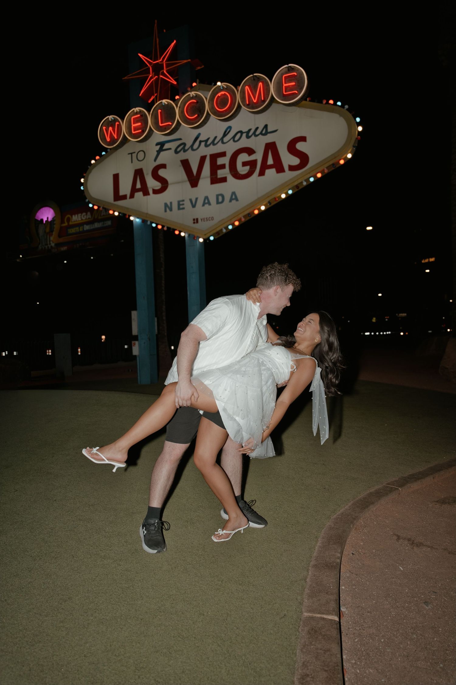 Man lifting woman in white dress for romantic photo at the Welcome to Fabulous Las Vegas sign at night.