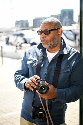 Middle-aged Black man in denim jacket and sunglasses holding a camera outdoors near a marina.