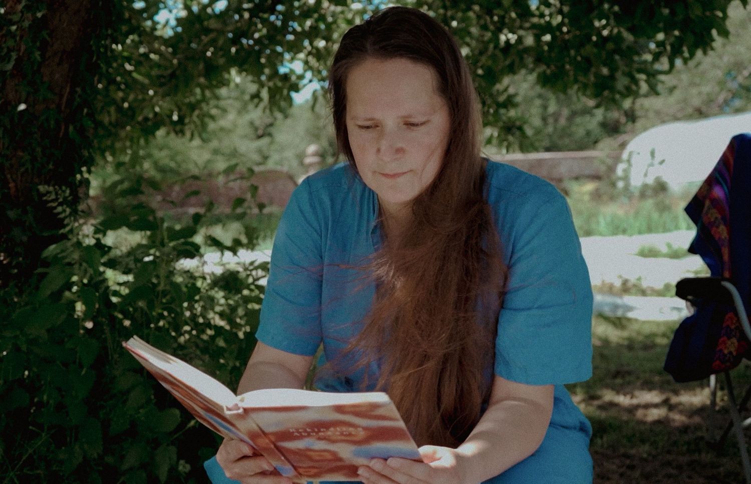 Still from a video. Female with long hair reading a colourful book outdoors under green trees on a sunny day.