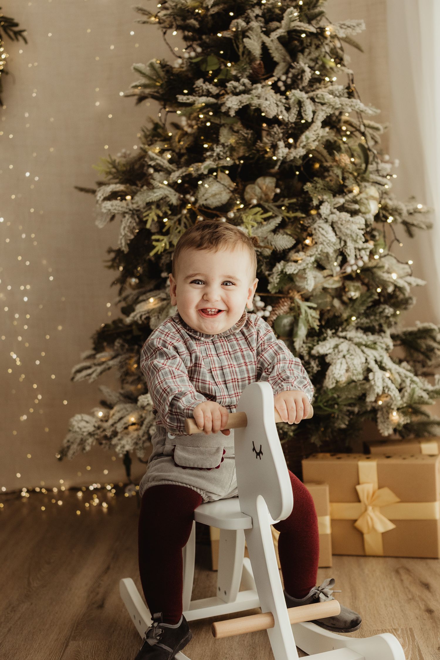 Adorable baby boy in plaid shirt holding a gold glitter Christmas ornament ball while sitting near a decorated tree.