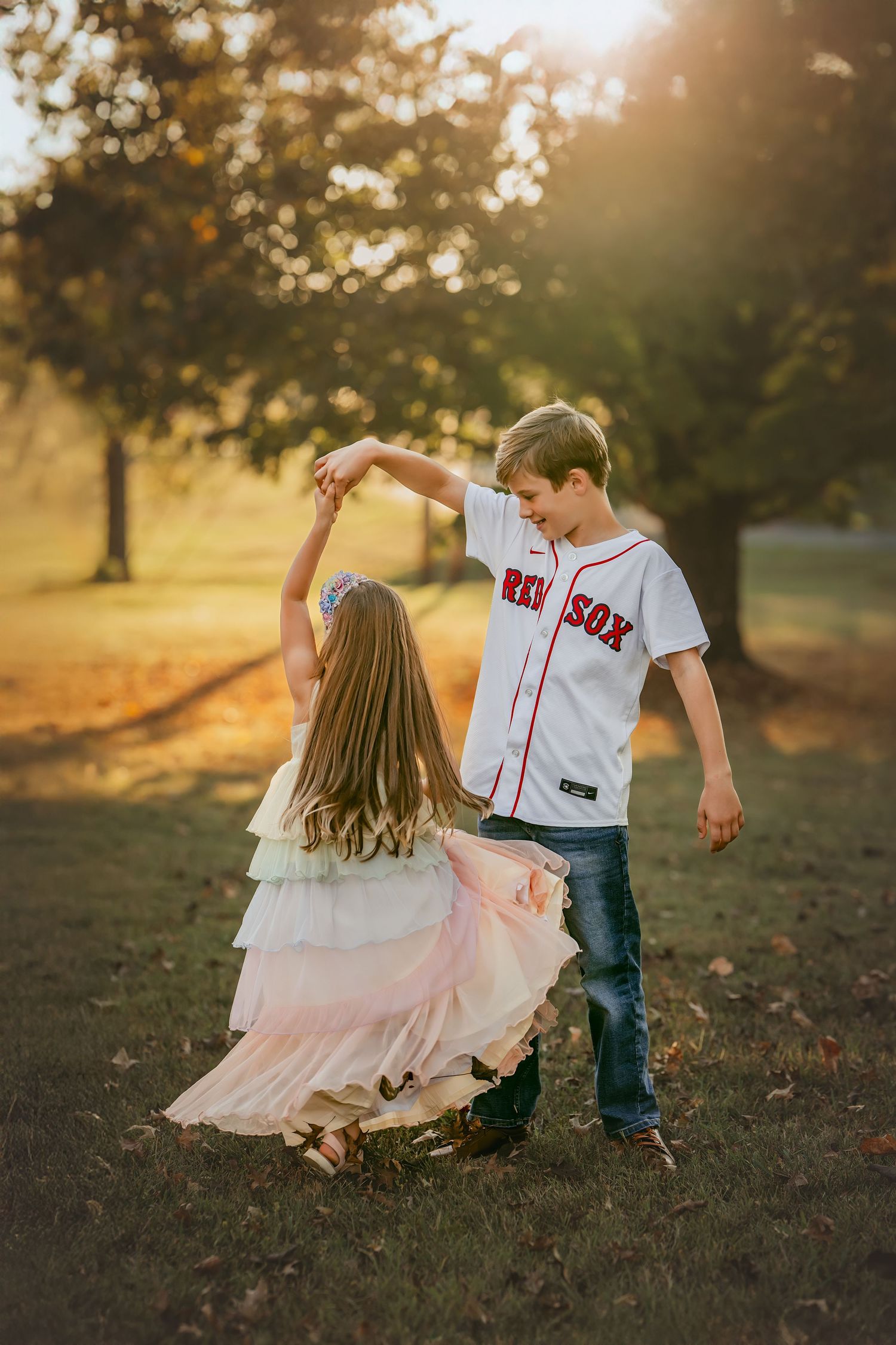 Two children dancing outdoors at golden hour, boy in Red Sox jersey twirling girl in flowing dress.