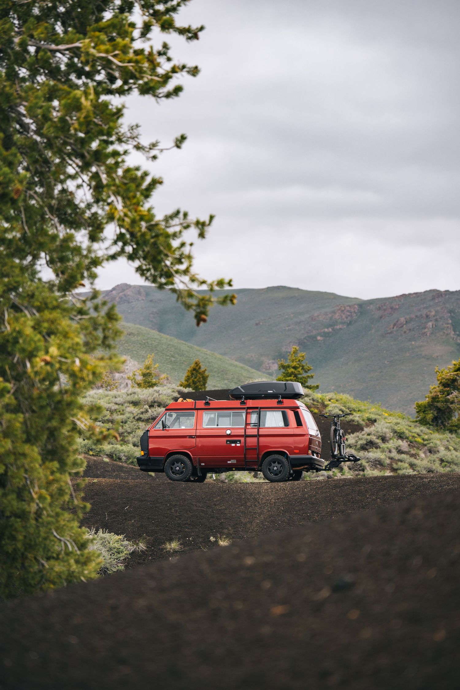 Red vintage van parked on hillside with mountains in background under cloudy sky.