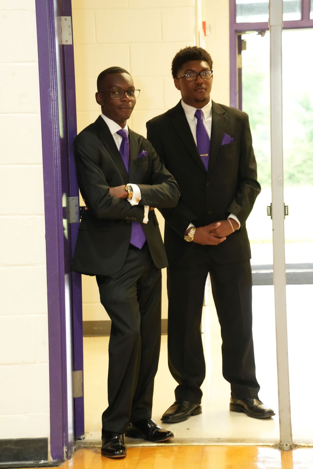 Two young men in matching black suits with purple ties and pocket squares stand in a doorway.