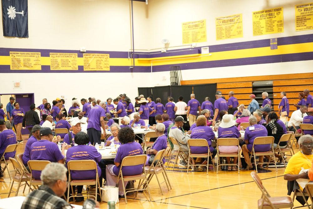 Large group gathering in a school gymnasium with people wearing matching purple t-shirts seated at round tables.