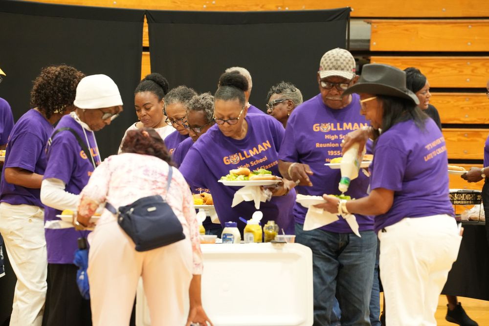 Group in matching purple Grillman t-shirts serve food at buffet table during gymnasium community event.