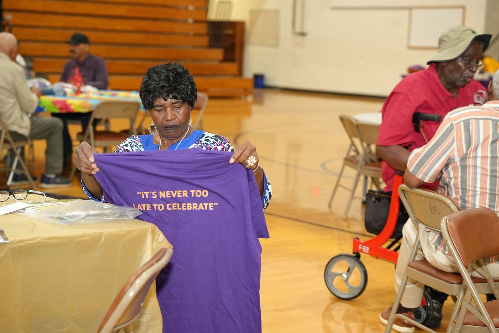 Woman holds up purple t-shirt reading It's Never Too Late to Celebrate at indoor community event in gymnasium.