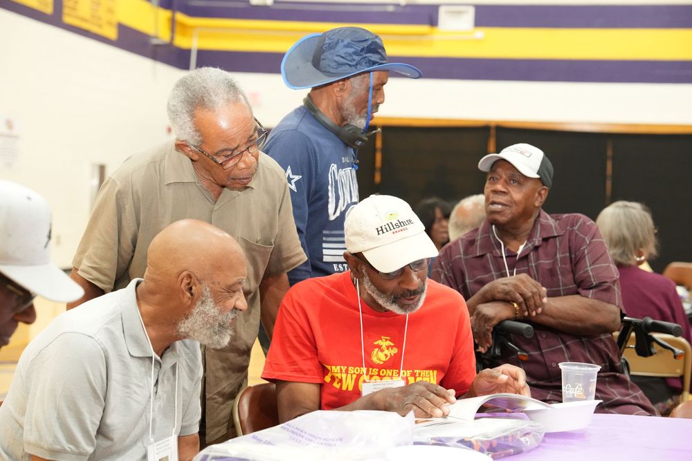 Group of elderly men gathered around a table at a community event, one signing documents in a gym.