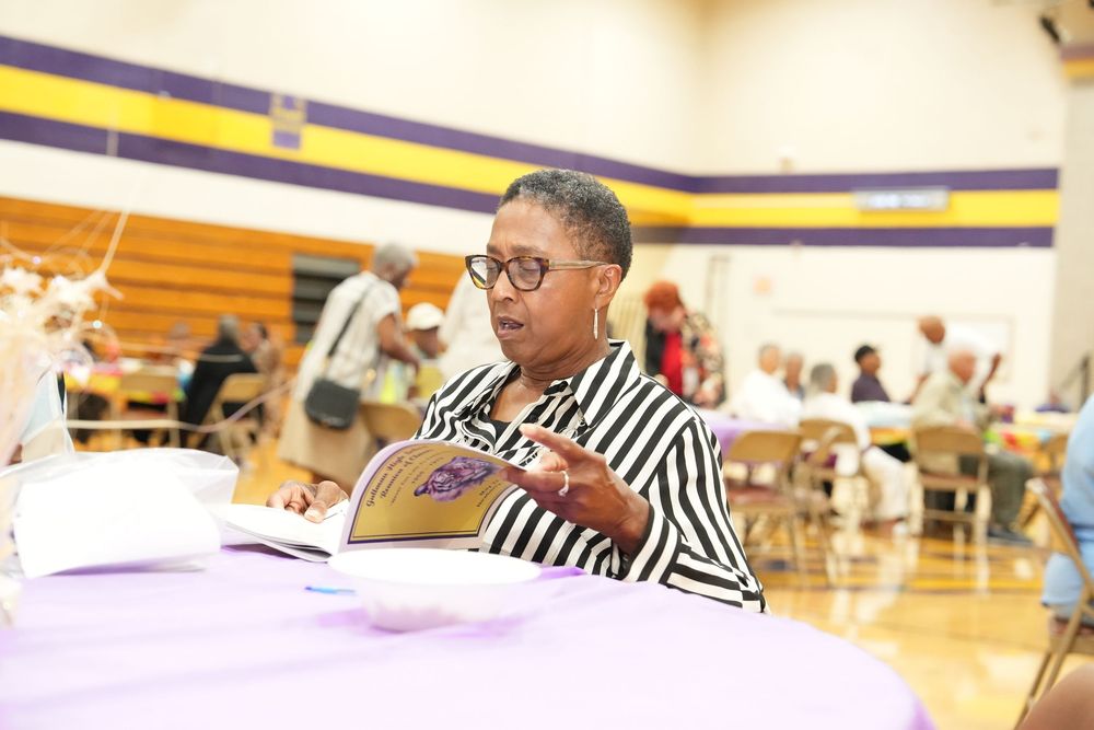 Woman wearing glasses reads book at purple-draped table during community gathering in school gymnasium.