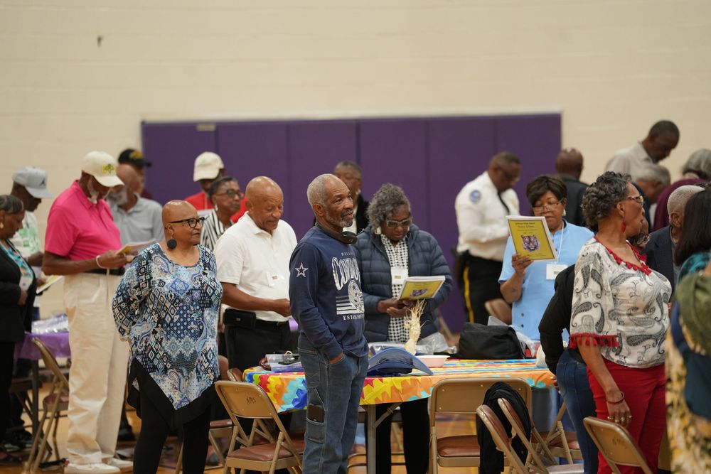 A diverse group of people browse tables at an indoor community event in a gymnasium with purple accents.