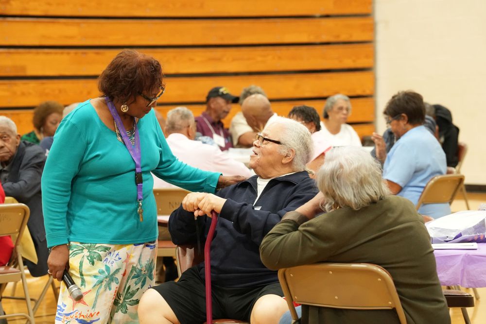 A woman in teal greets seated guests at a lively community gathering inside a gymnasium.