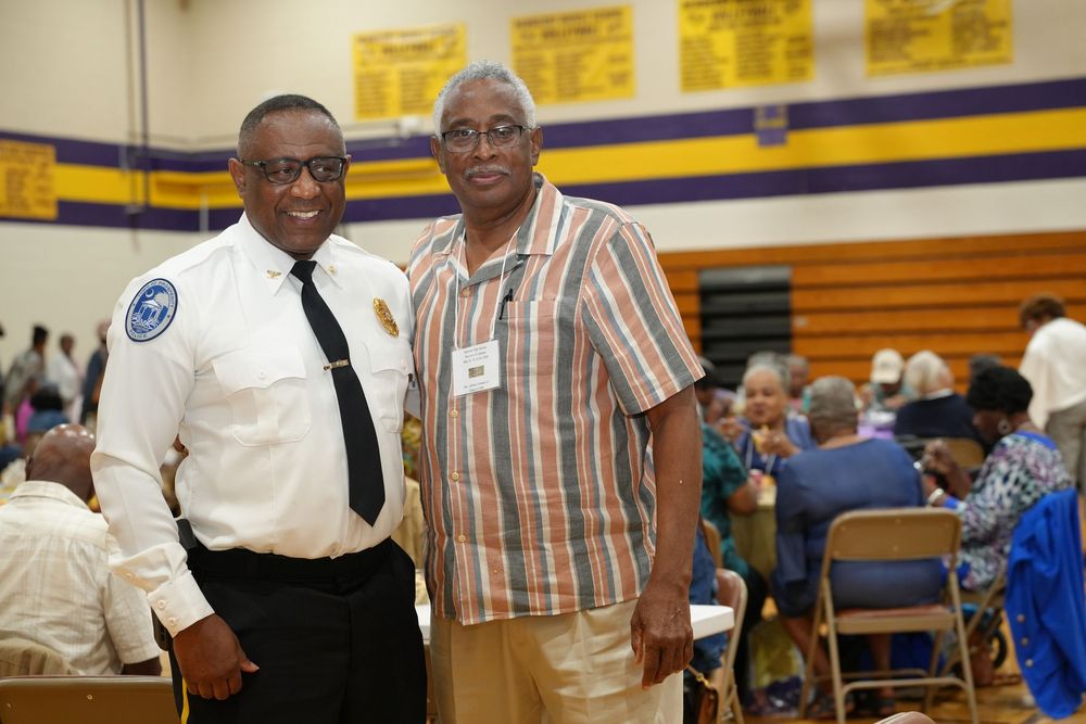 Two men pose together smiling in a school gymnasium during a community event, one wearing a security uniform.