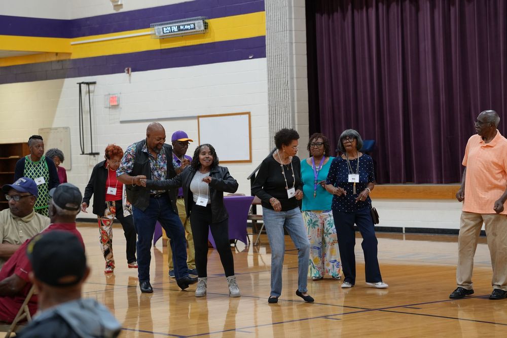 Group of people line dancing in a school gymnasium with purple and gold decorations during a community event.