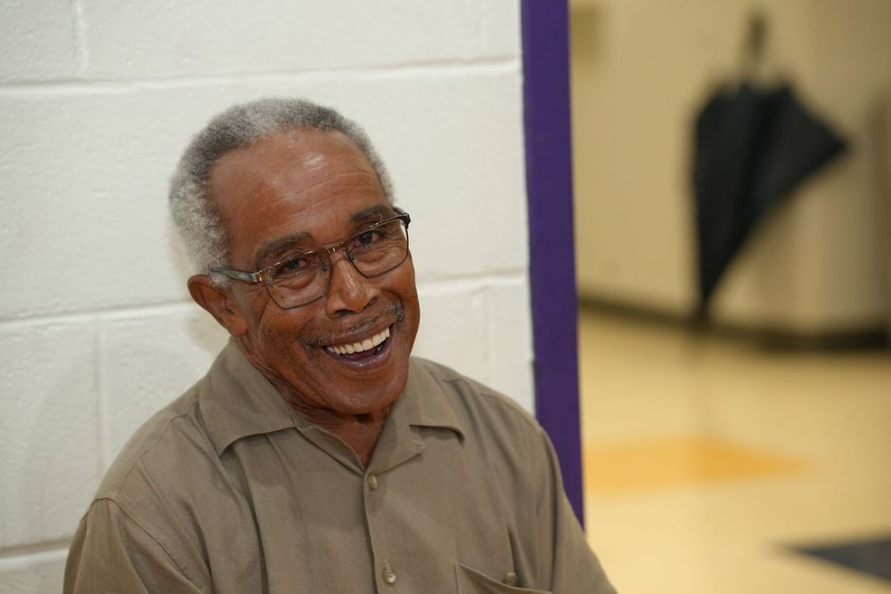 Smiling elderly man with glasses sitting in a gymnasium at a community gathering event.