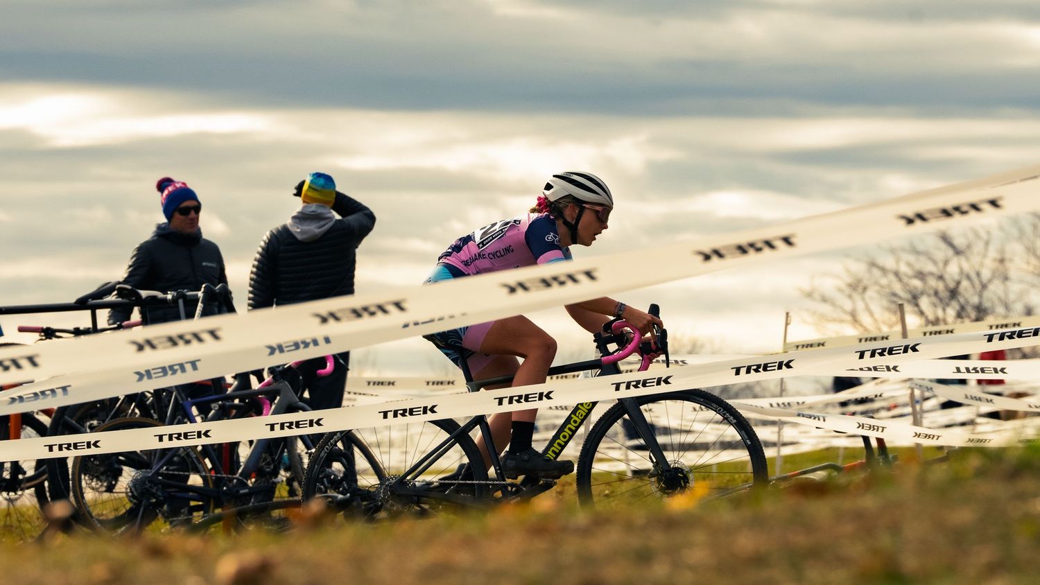 A cyclocross racer in a pink kit rides a course marked with TREK tape under a gray autumn sky, spectators and bikes lining the sideline.