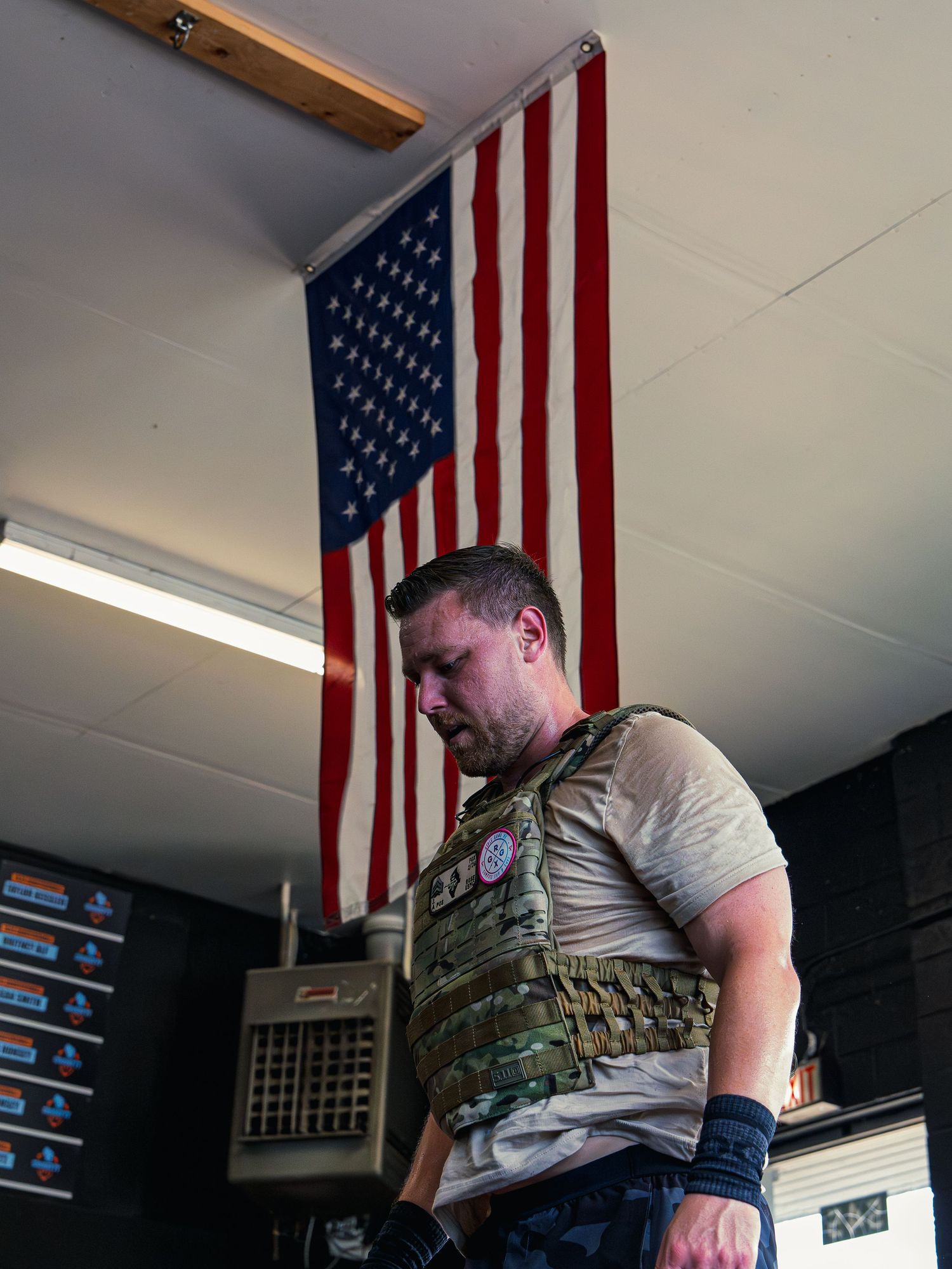 Sweat-soaked CrossFit athlete in a camo weight vest catches his breath during Murph, an American flag hanging on the wall behind him.