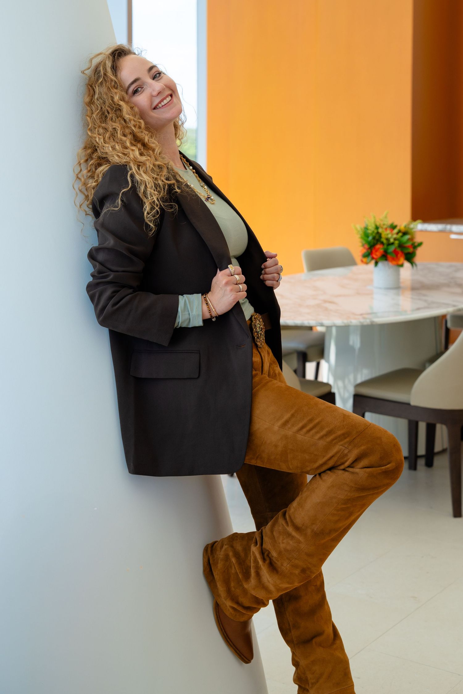 Smiling woman with curly blonde hair wearing a black blazer, brown suede boots, and tan pants leaning against a white wall.