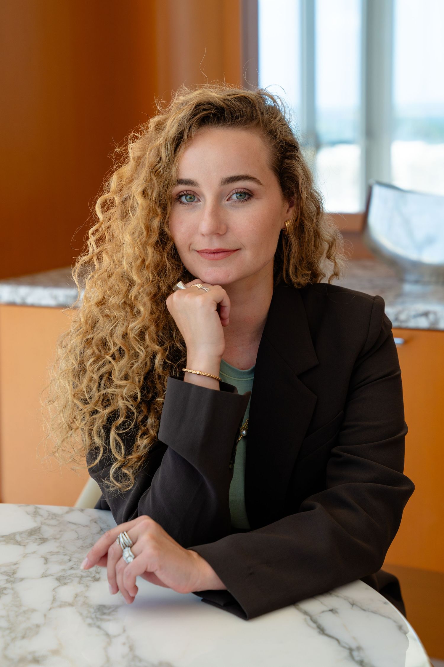 Woman with curly blonde hair in black blazer poses thoughtfully at marble table in modern interior.