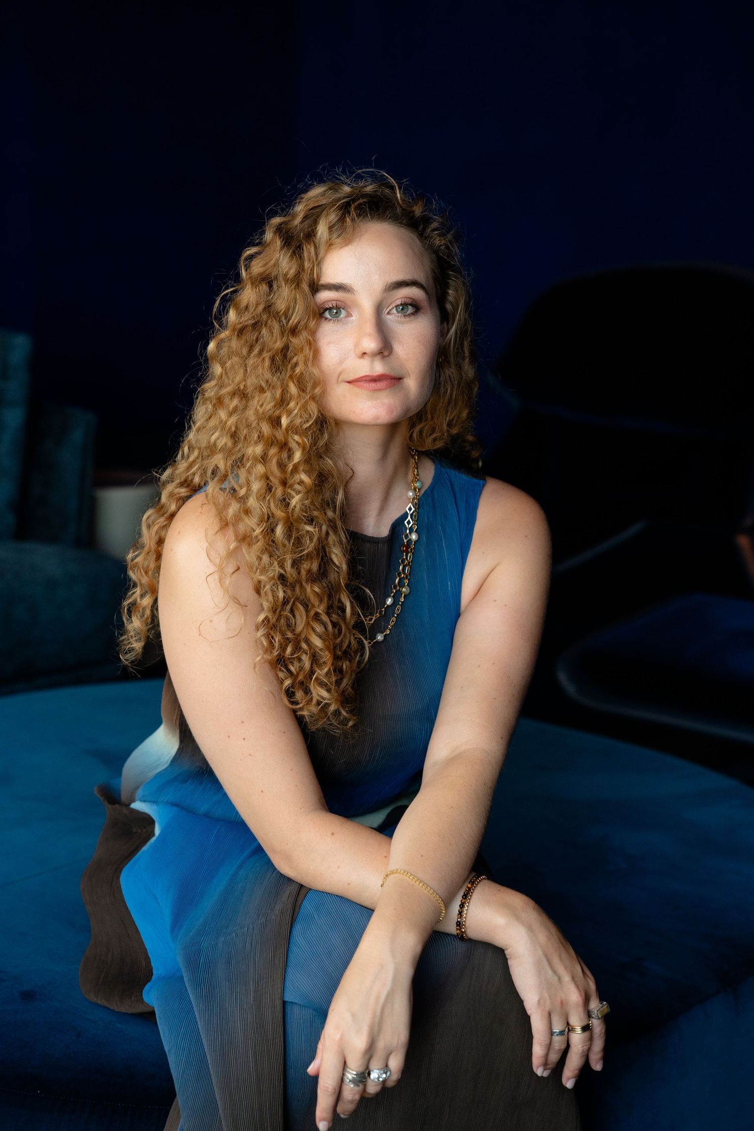 A woman with long curly auburn hair wearing a blue sleeveless dress sits posed against a dark blue background.