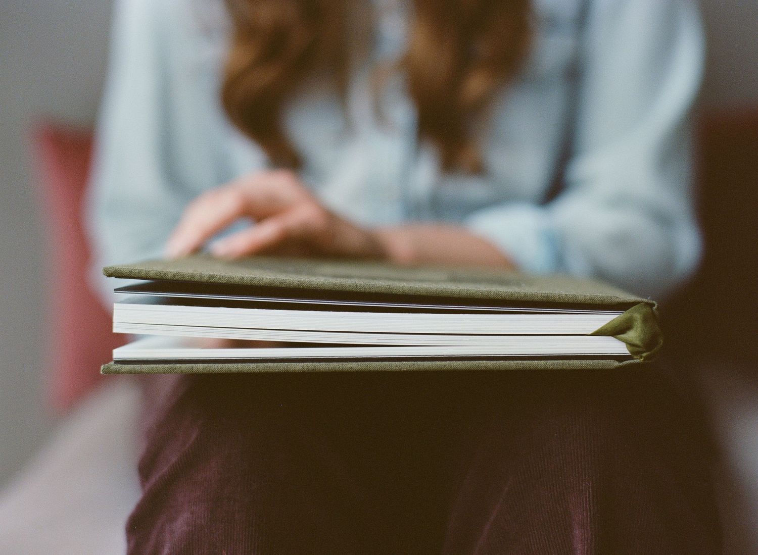 A young woman with red hair reads a thick hardcover book with a green cover while sitting down.