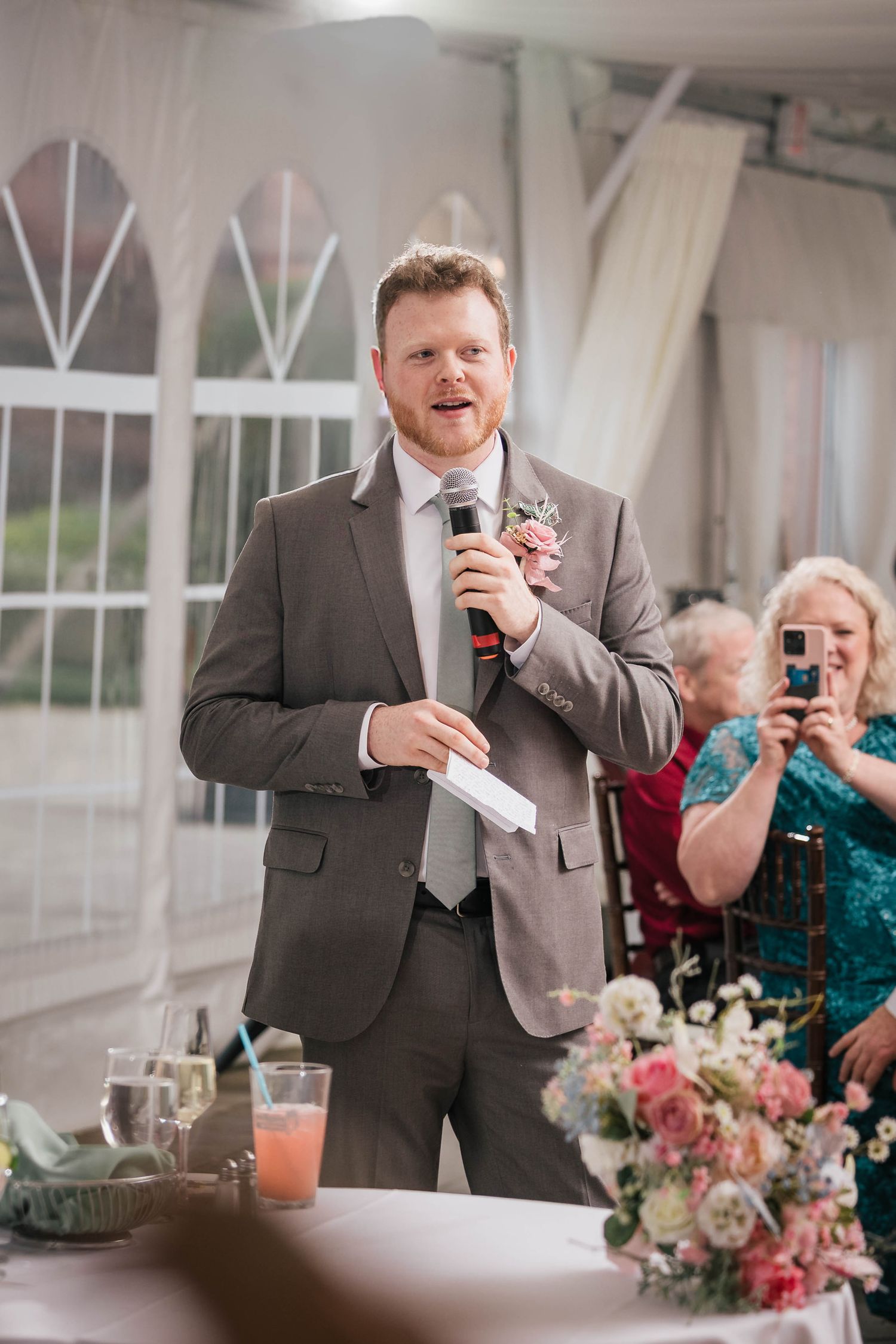 Groomsman giving a speech from a handwritten card, guests smiling in the background.
