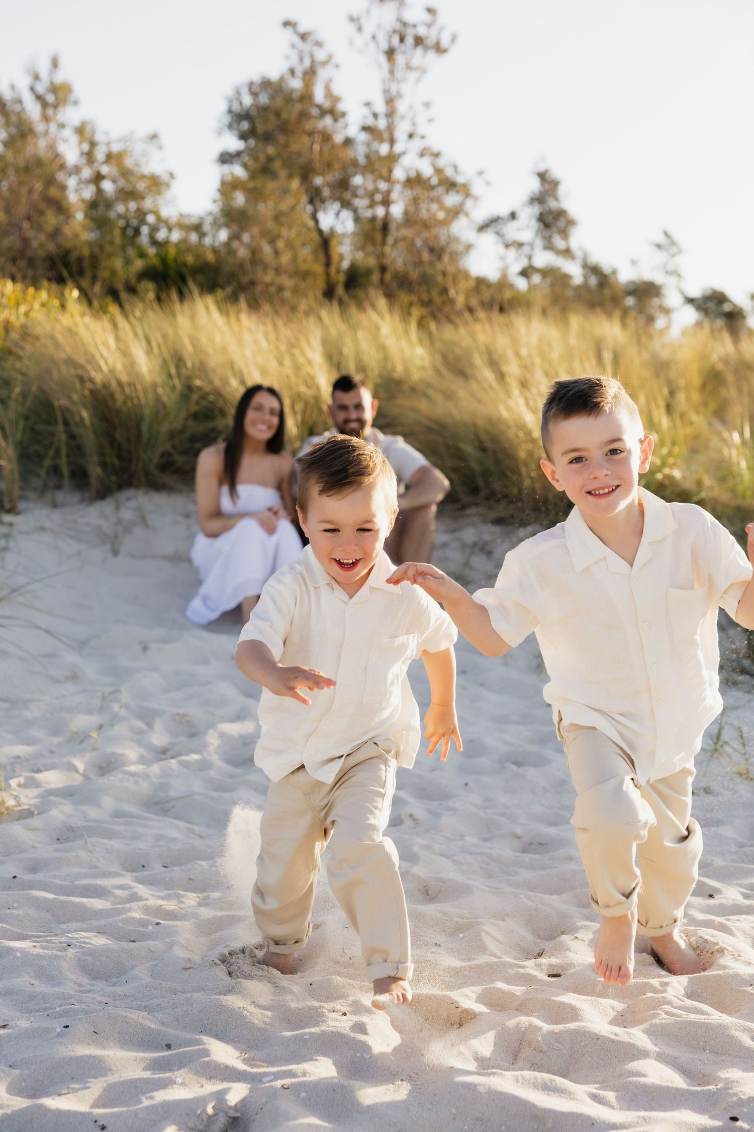 Two young boys in white shirts run barefoot on sandy beach dunes while their parents relax in the background.