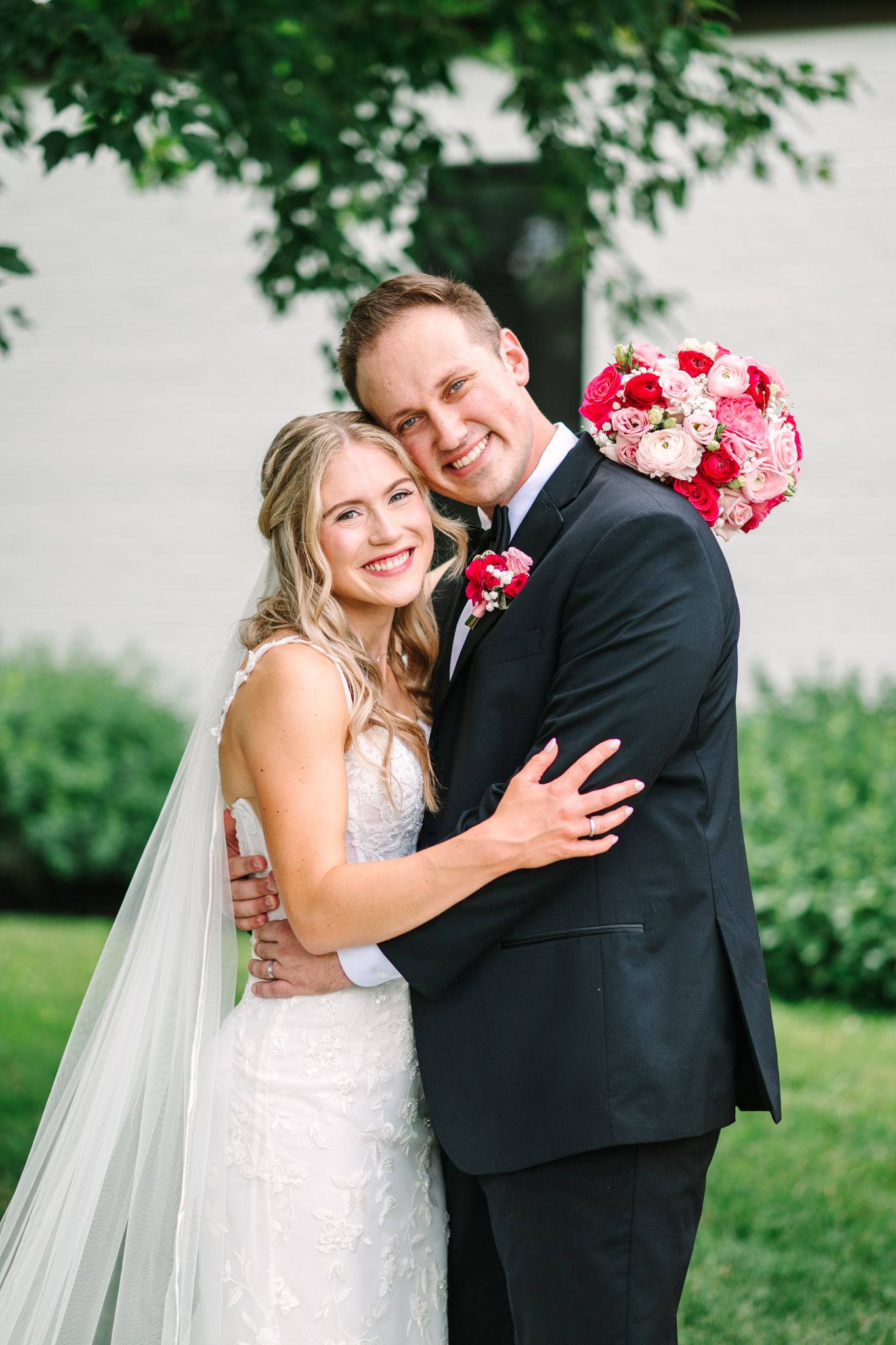 Smiling bride and groom embrace outdoors, bride in lace gown with veil, groom in black tux, holding pink bouquet.