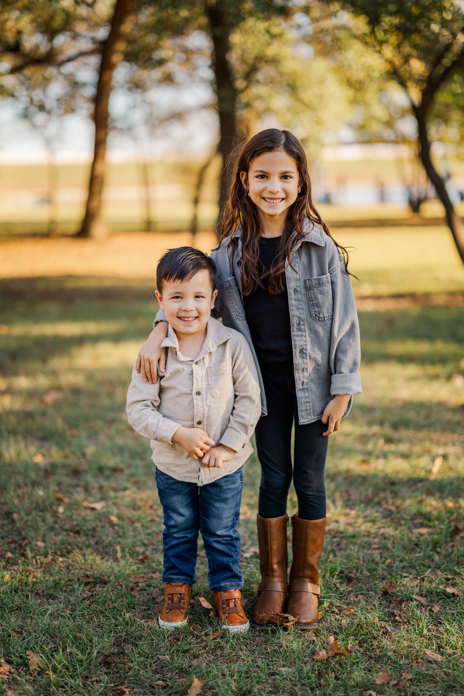 Two smiling children posing outdoors in a park during autumn, wearing casual jackets and jeans.