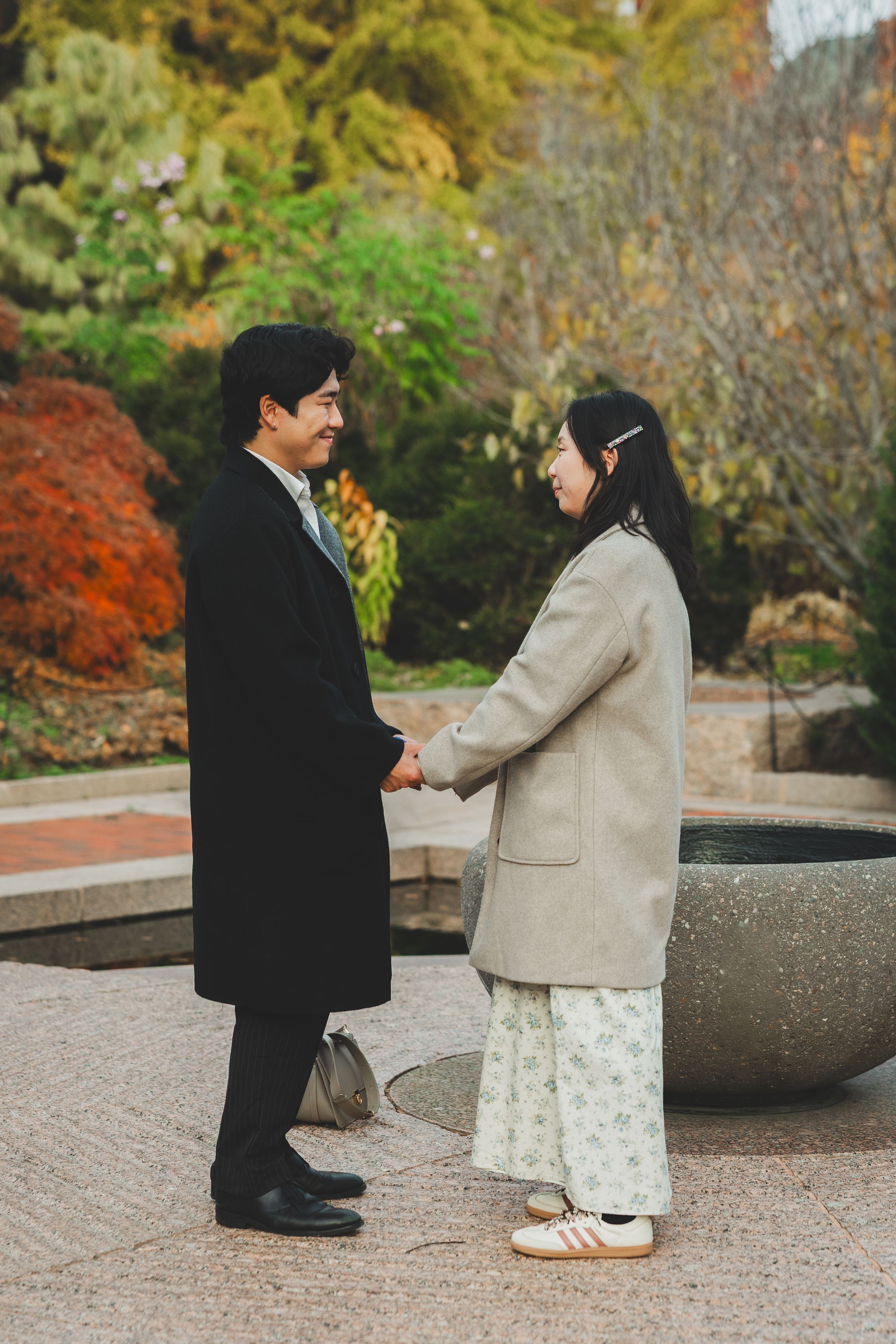 A couple holds hands and smiles at each other in an autumn garden, dressed in elegant coats.