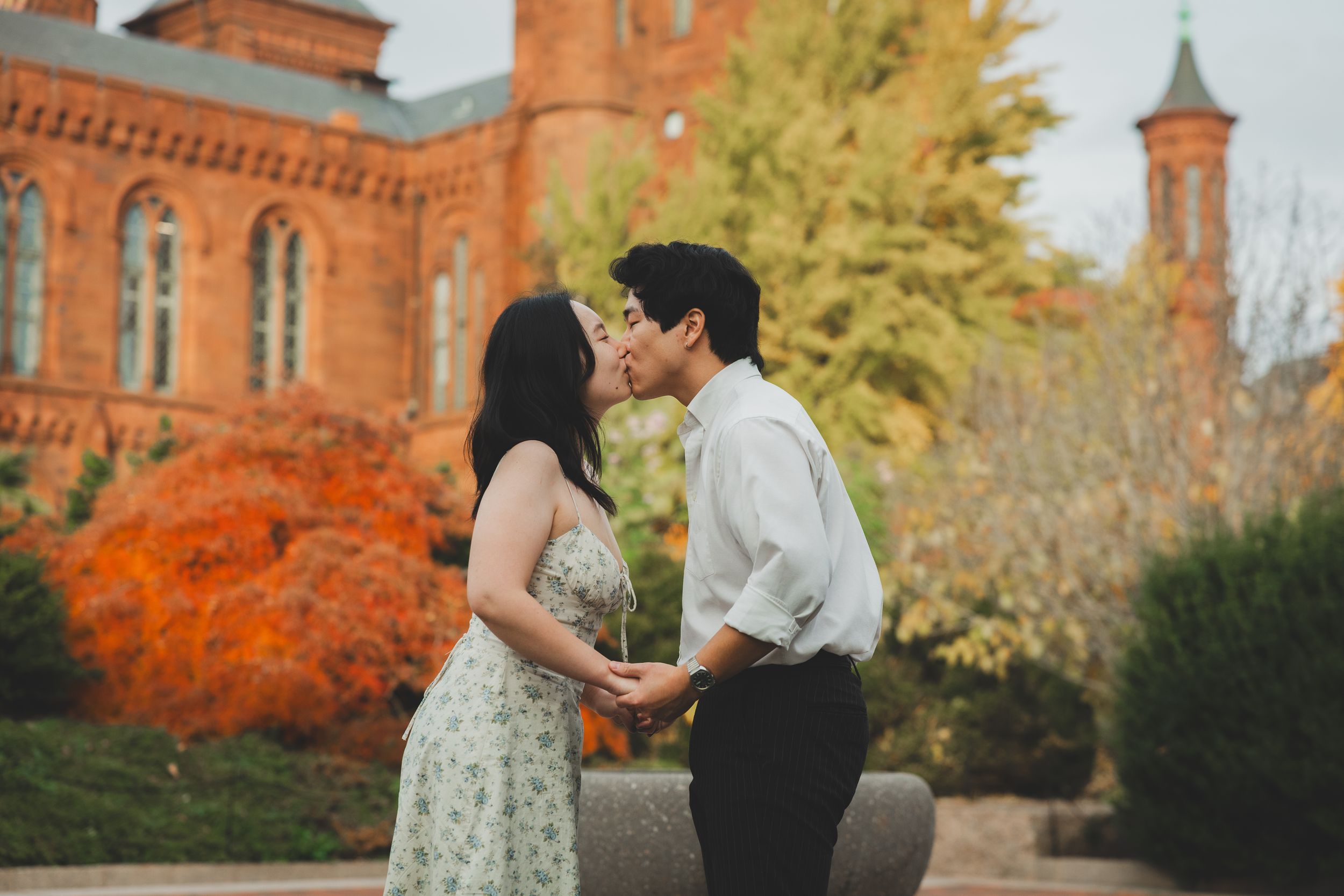 Couple sharing a romantic kiss outdoors near a red brick building surrounded by vibrant autumn foliage.
