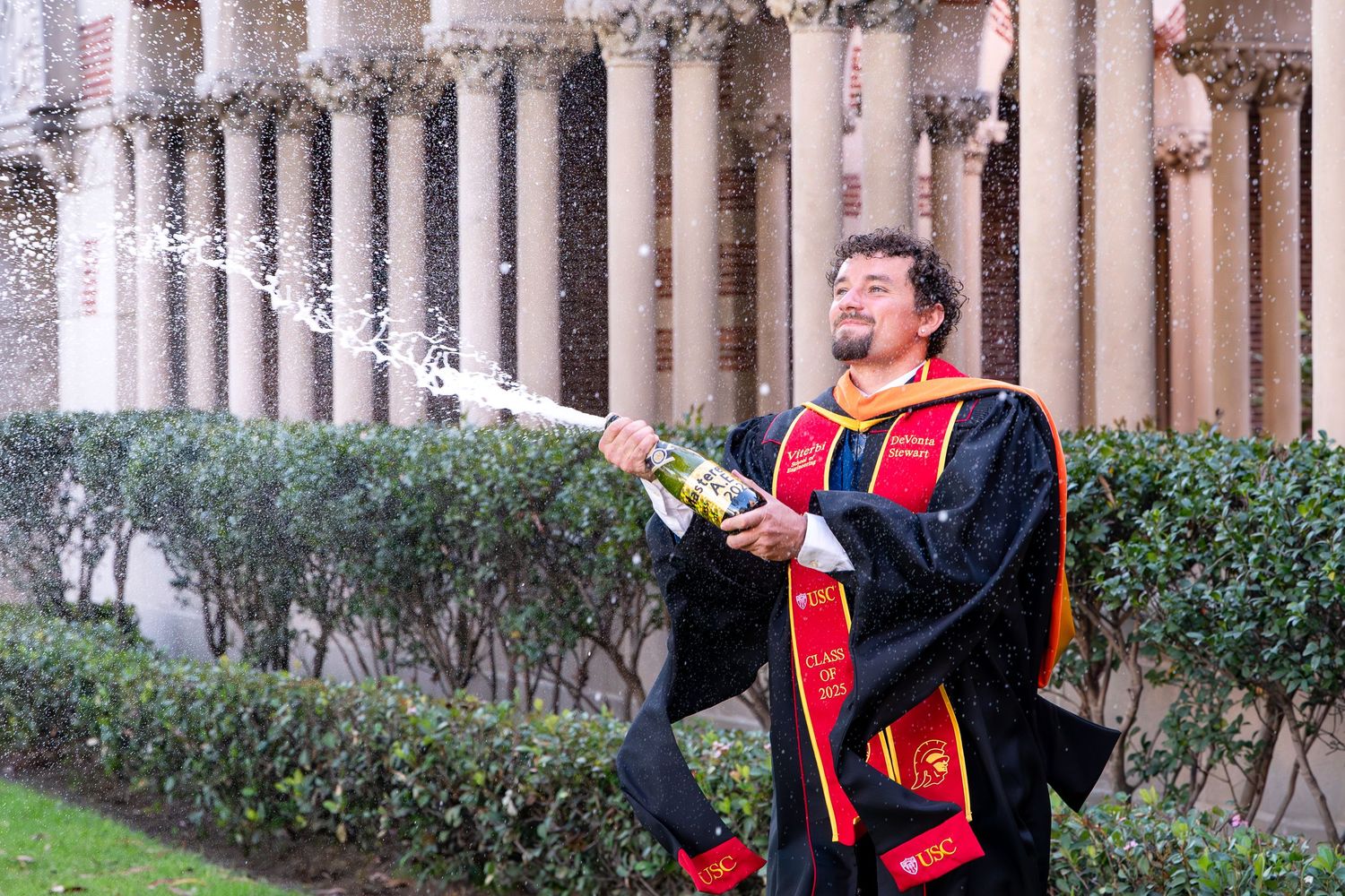 Graduate in academic regalia sprays champagne outdoors near stone columns celebrating graduation day.