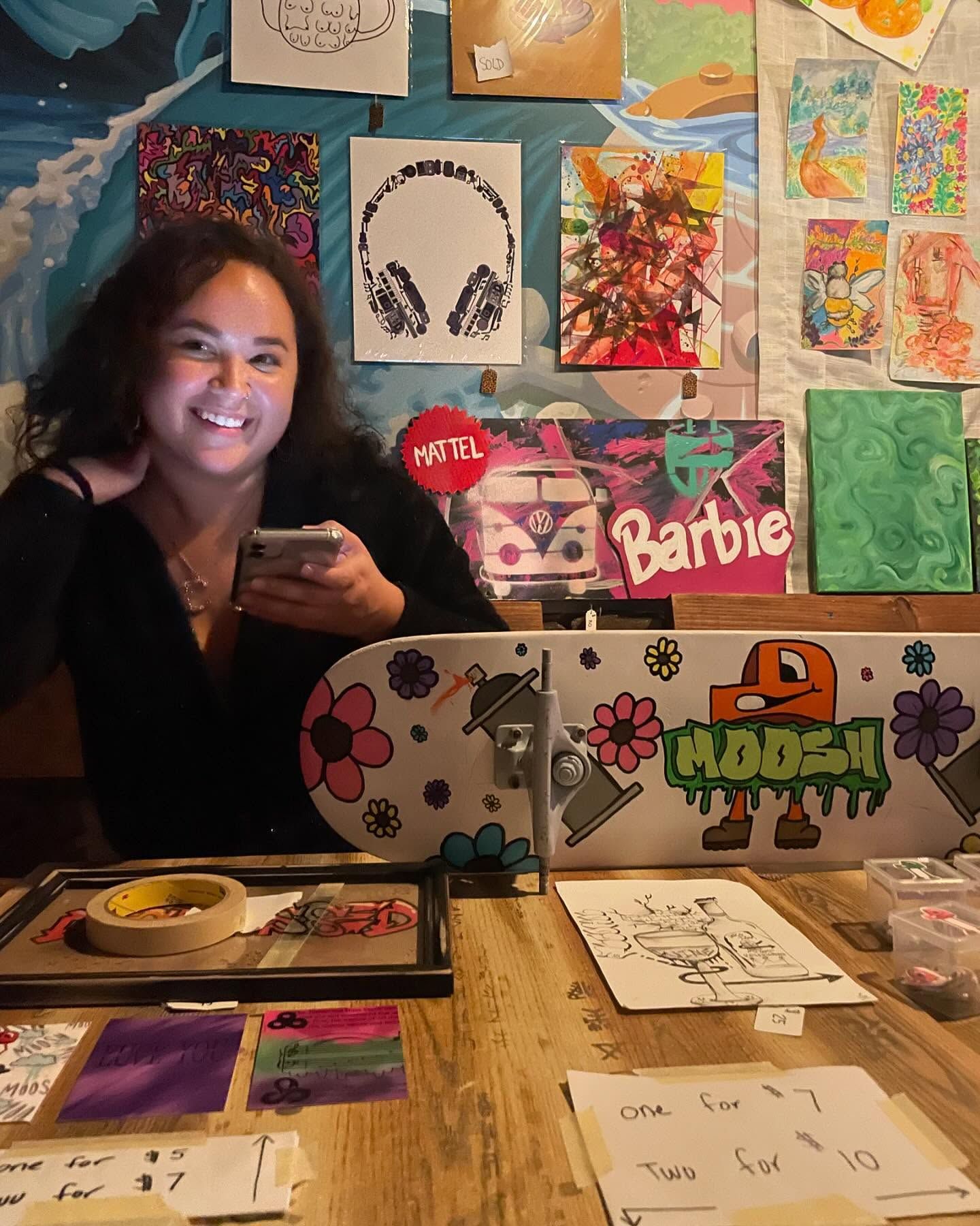 Artist smiling behind a table displaying custom painted skateboards and colorful artwork at an indoor art market.