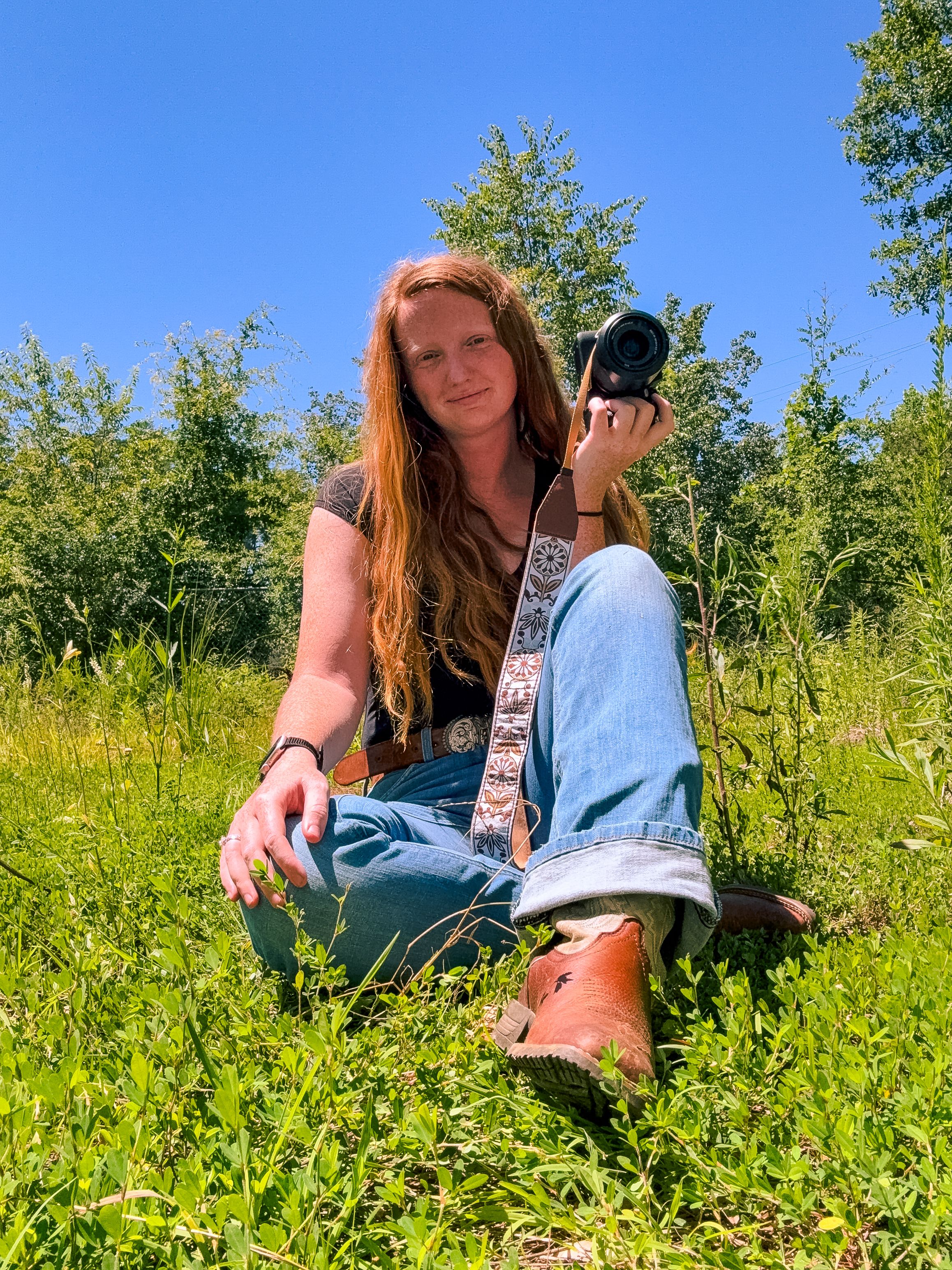 Redhead woman sitting in green field holding camera on sunny blue sky day.