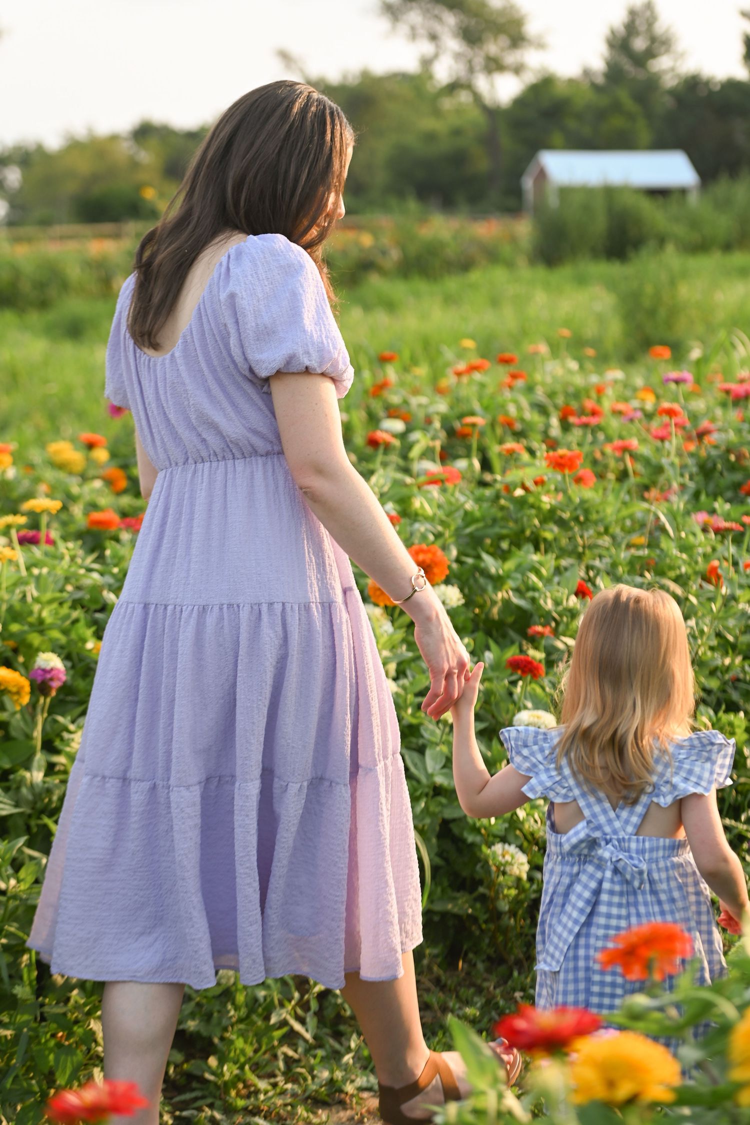 A mother and young daughter in floral dresses walk hand-in-hand through a vibrant zinnia flower field.