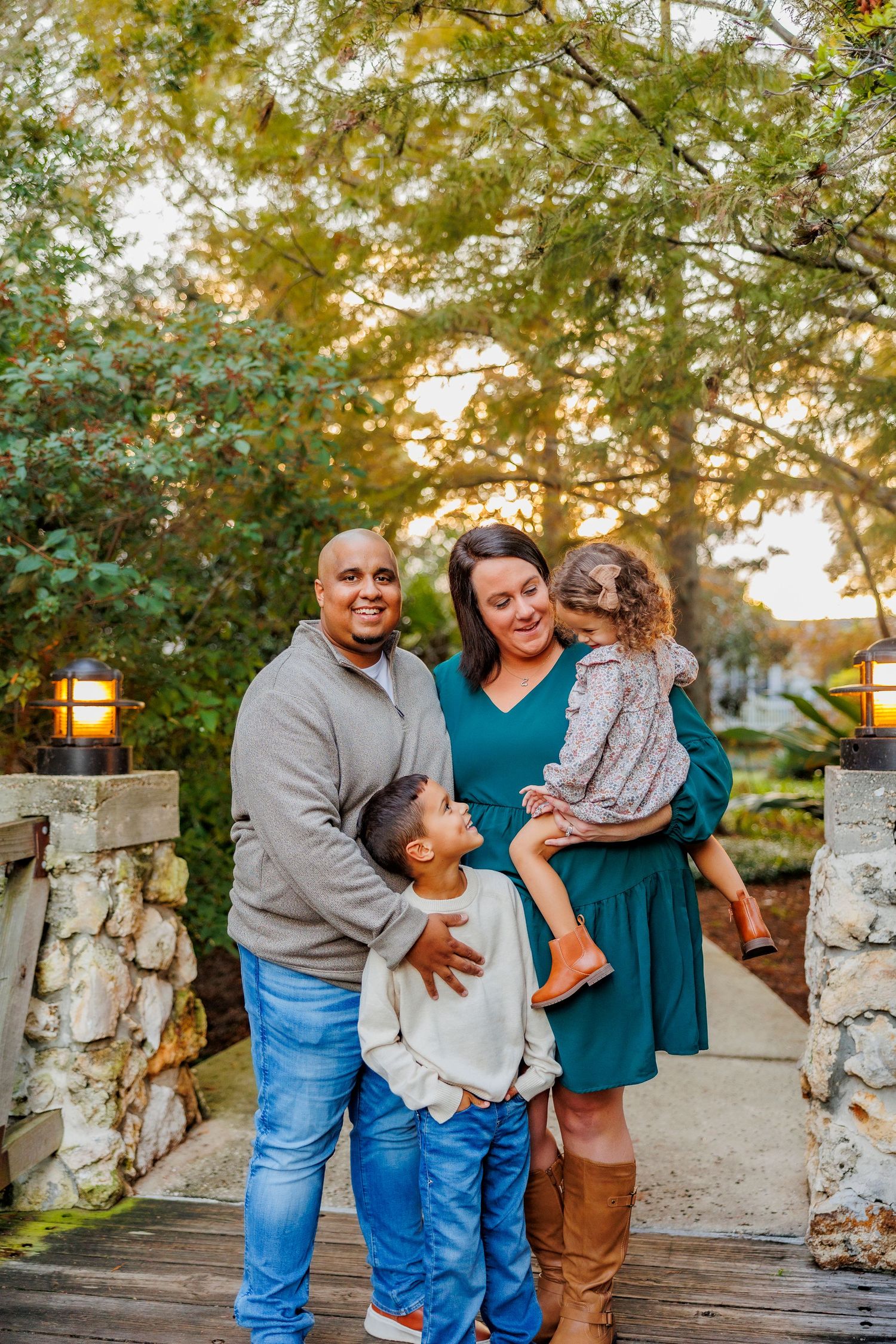 A happy family of four poses outdoors on a wooden path surrounded by lush trees and warm garden lights at sunset.
