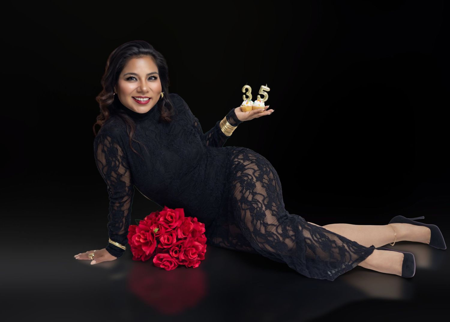 Woman in black lace jumpsuit poses on floor holding gold award with red roses bouquet on dark background.