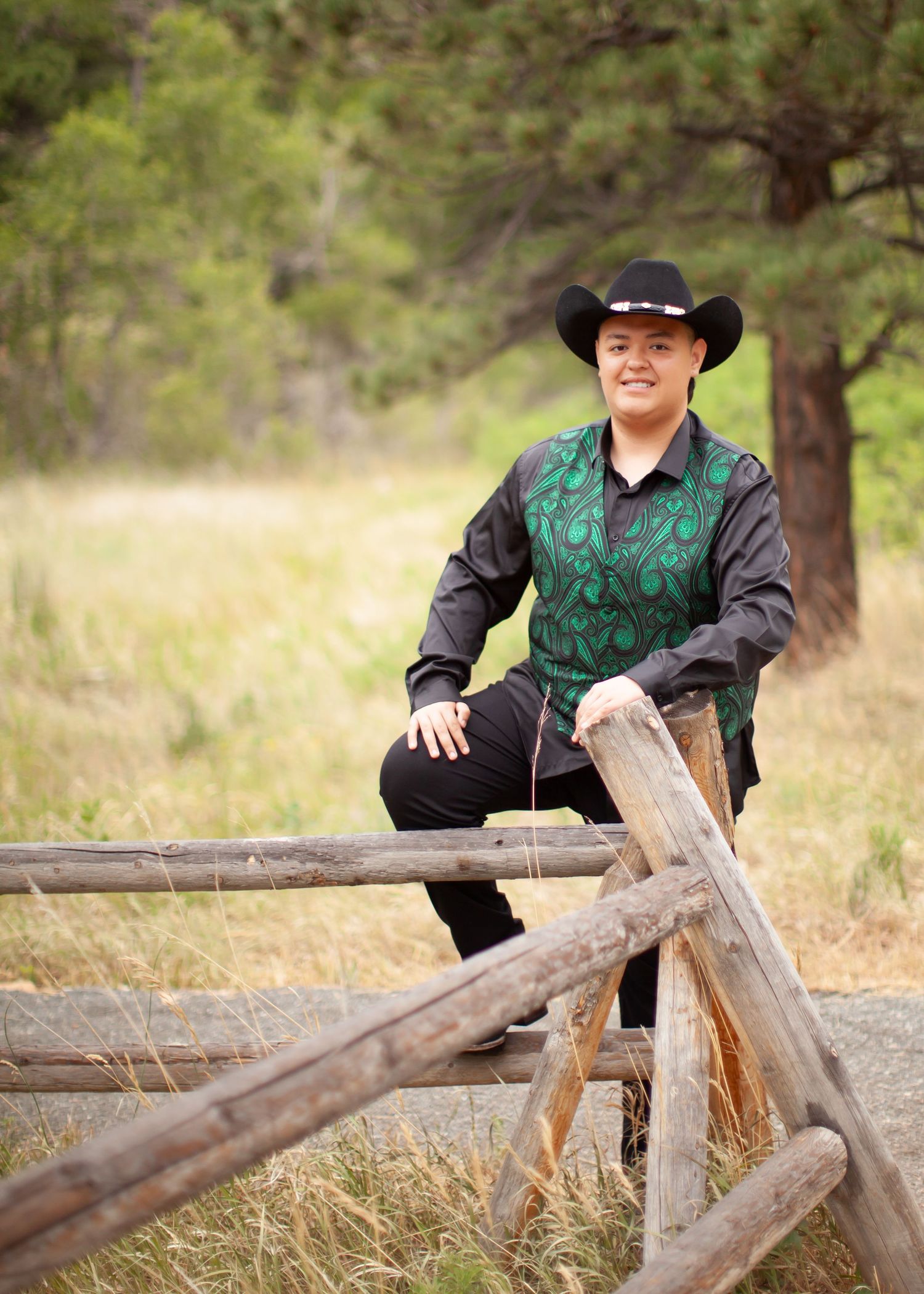 Person in black cowboy hat and green vest leaning on rustic wooden fence in a natural outdoor setting.
