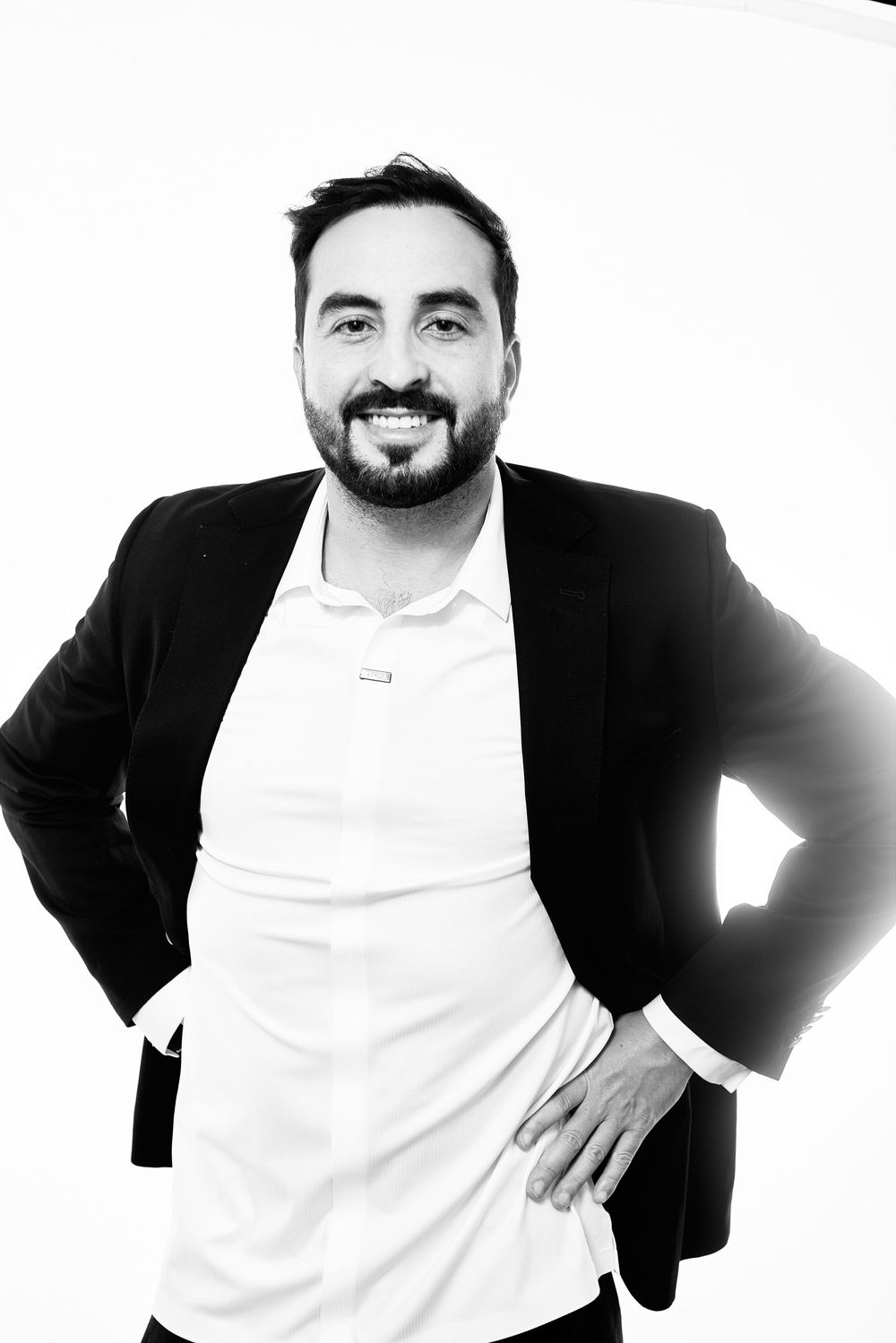 Black and white portrait of a smiling bearded man in a blazer and white shirt, hands on hips.