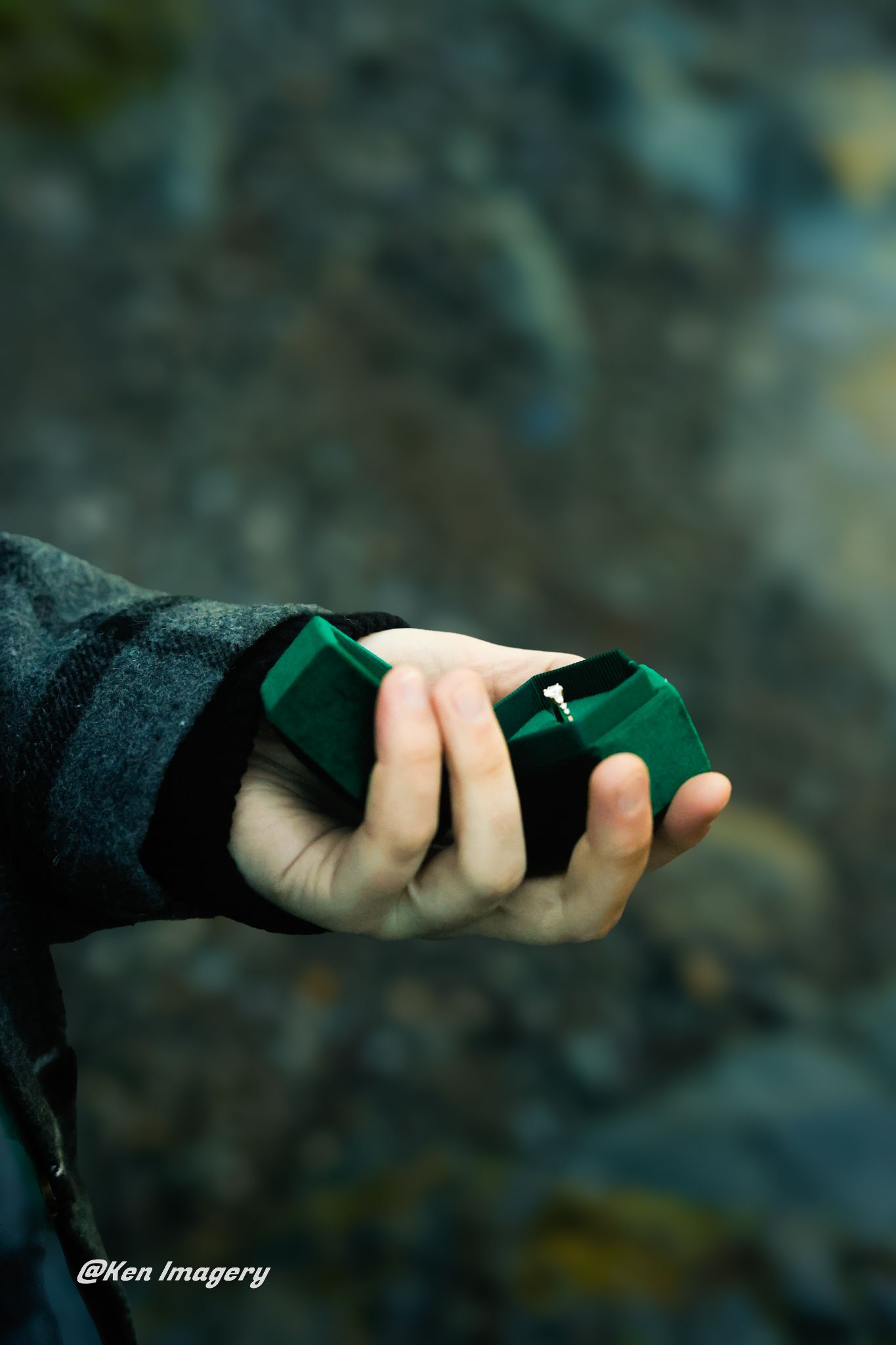 Hand holding open green velvet ring box with diamond engagement ring outdoors against rocky background.