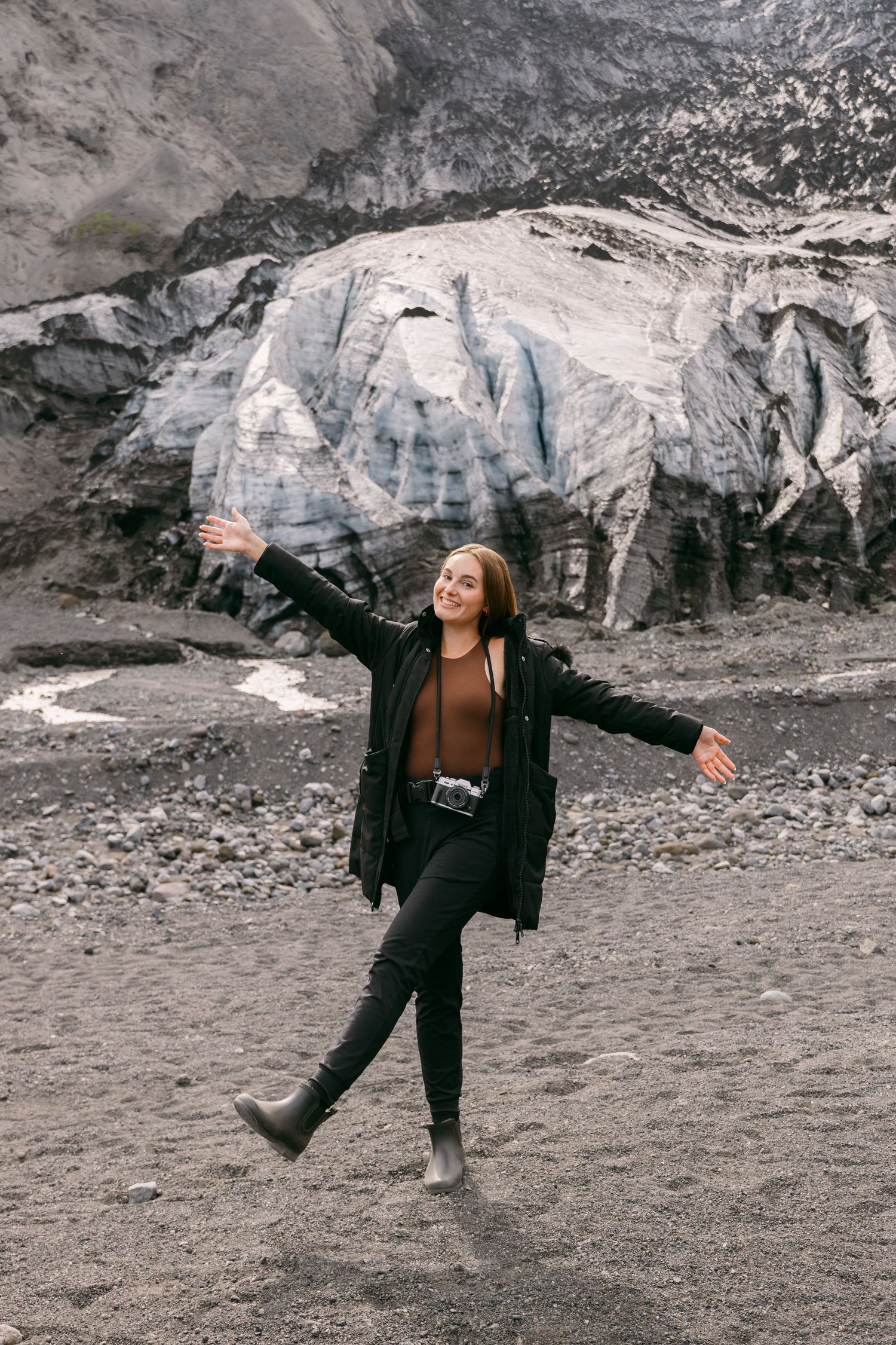 A joyful woman in black jacket poses with arms outstretched near a glacier on a rocky terrain.