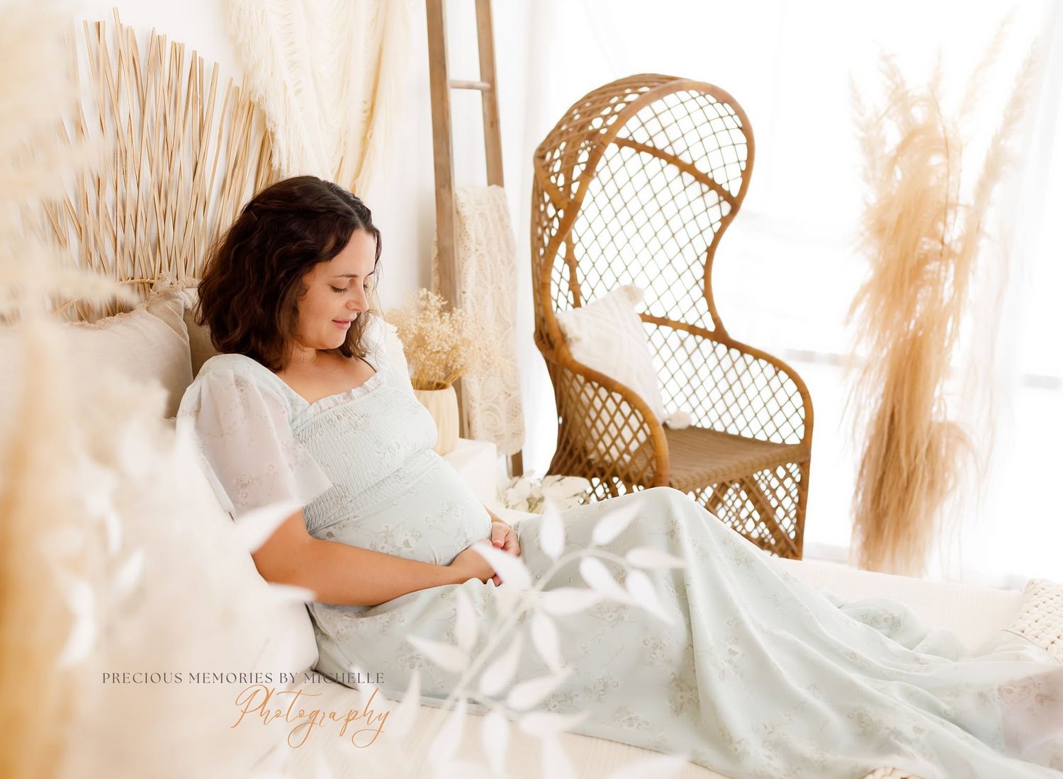 Pregnant woman in light blue floral dress seated on vintage chair, gazing down at baby bump in soft natural light.
