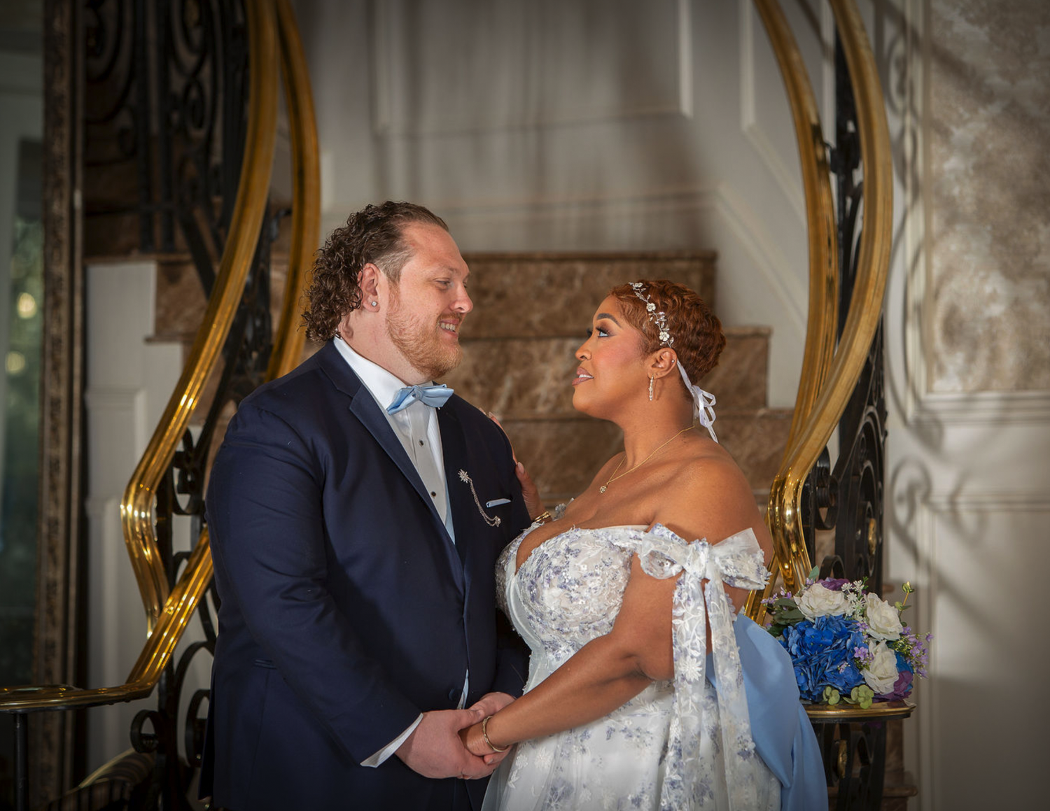 Bride and groom share a romantic kiss at elegant staircase, bride in floral white gown with blue sash, groom in navy suit.